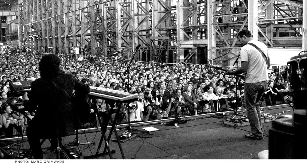 A black and white photograph of two people on stage in front of a large crowd playing guitar.