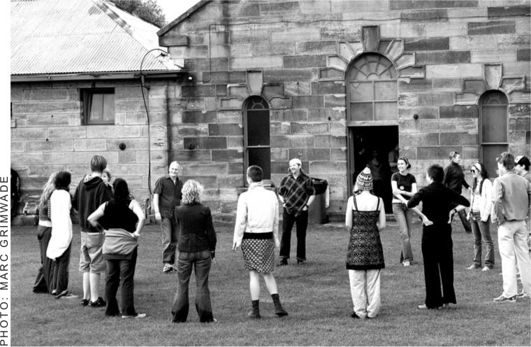 A black and white photograph of a group of men and women standing in a large circle in front of a sandstone building.
