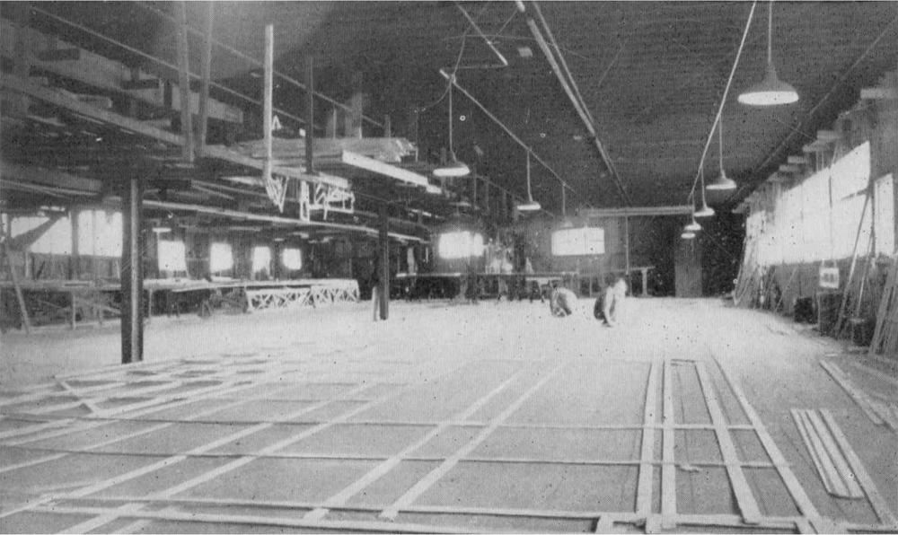 A black and white photograph of the inside of a loft with people working on the floor and low hanging lights around the building.