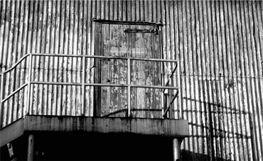 A black and white photograph of a door on the edge of a tin building.