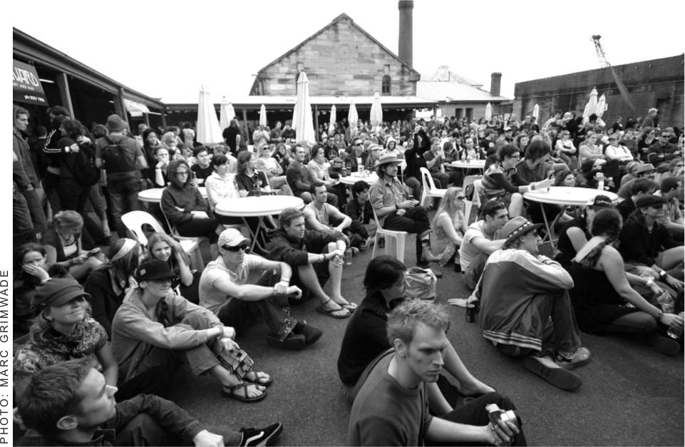 A black and white photograph of men and women sitting and standing in the crowd of a performance at Cockatoo Island.