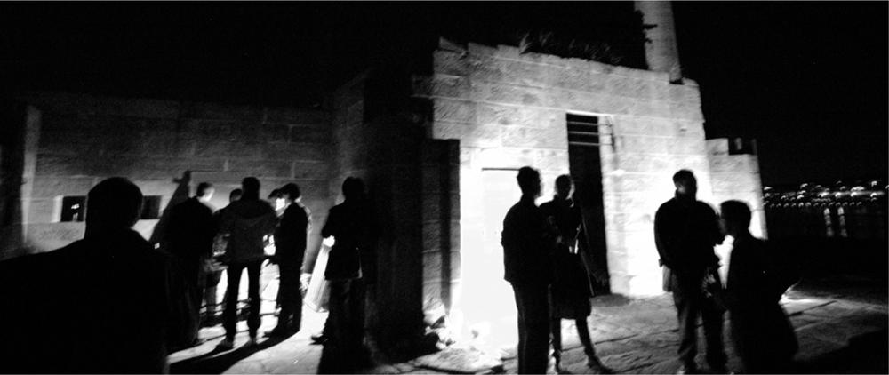 A black and white photograph of men and women in front of the lights of an old sandstone building at night.