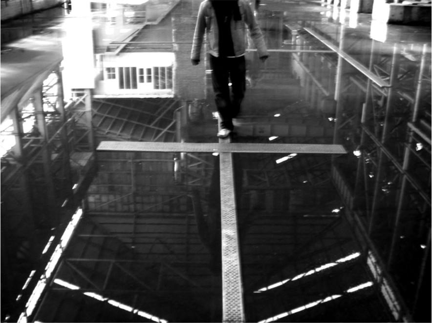 A black and white photograph of a man walking on an exposed beam in a room with water on the floor.