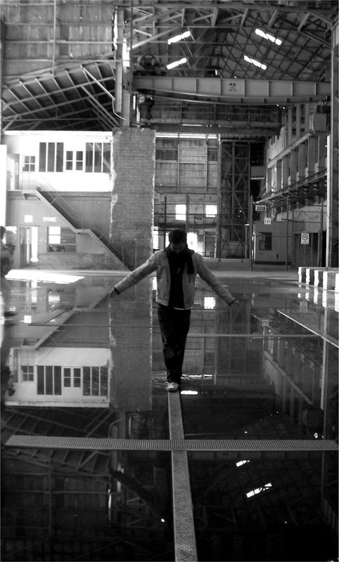 A black and white photograph of a man walking on beams in a large building with water on the floor.