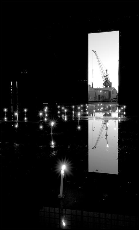 A black and white of a large room with a light installation and lights on the floor. There is a crane through a large door in the background.
