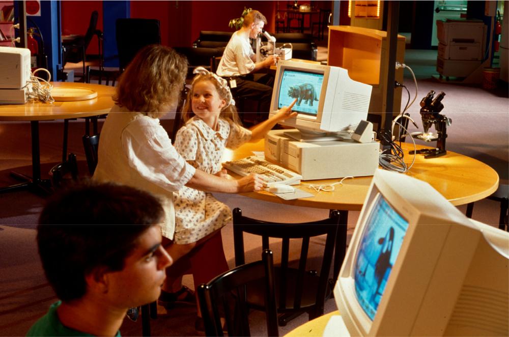 A photograph of kids using computers with a young girl sitting on a woman's lap and looking at animals on the computer.