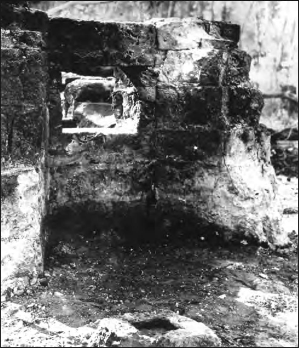 A black and white photograph of the smithy's remains. It has a curved wall with a square window. On the ground is a square brick.