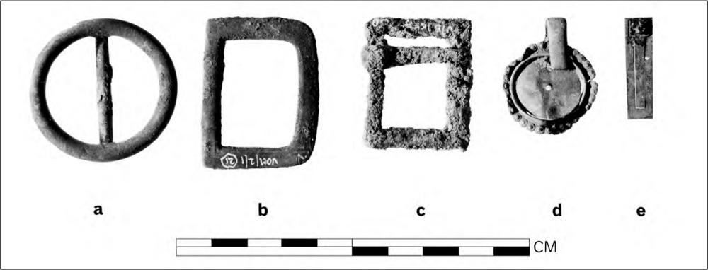 A black and white photograph of two brass belt buckles - one round and one square, a square iron belt buckle, a crass round brooch or cameo holder and a slim rectangular harmonica or accordion reed.