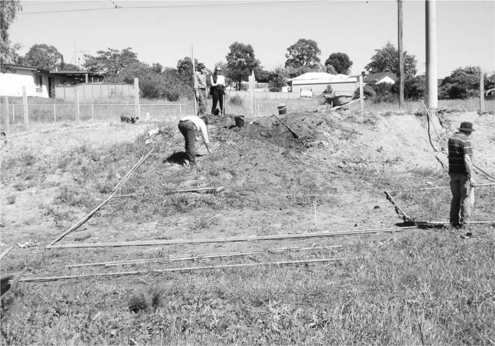 A black and white photo of a grass field with a dirt excavation site. There is a man bending over in the ground and two people standing on the top of a small hill. There is a man to the right of the image facing away from the camera.