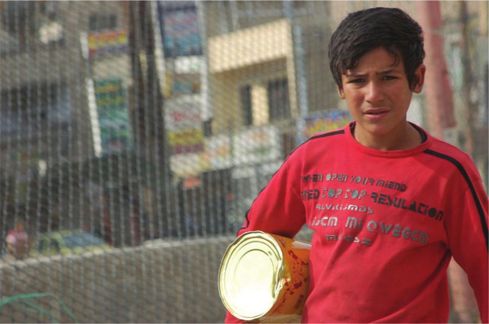 Photograph of a young boy in a red shirt, holding a tin can and standing in front of a wire fence.