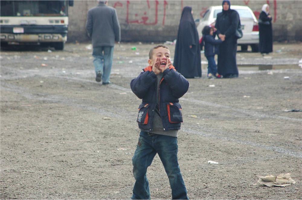 Photograph of a young boy standing in a bus depot. He wears blue jeans and a navy jacket, and holds his hand up to cup his mouth.