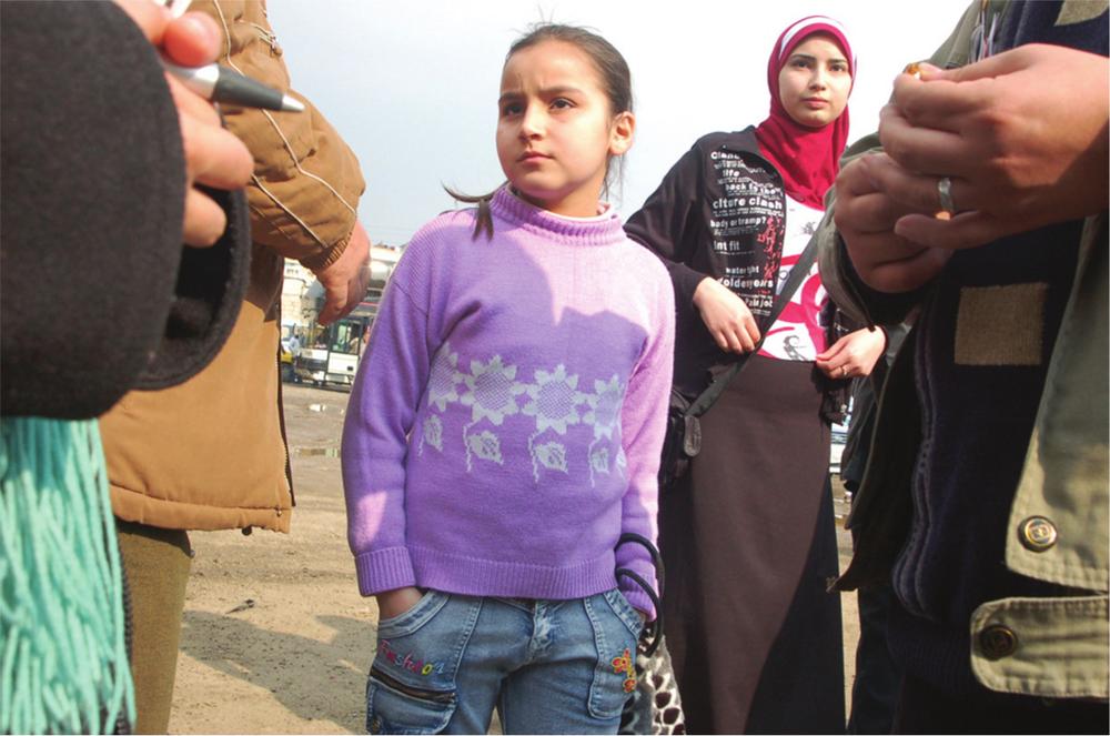 Photograph of a young girl wearing a purple sweater and blue jeans. An older girl stands behind her, and the hands and torso of three other adults are visible at the edges of the photograph.