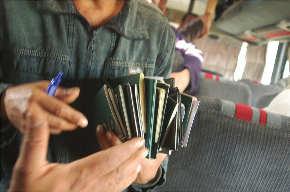 Photograph of a man's hands, holding a large pile of passports and a blue pen. Bus seats are visible in the background.