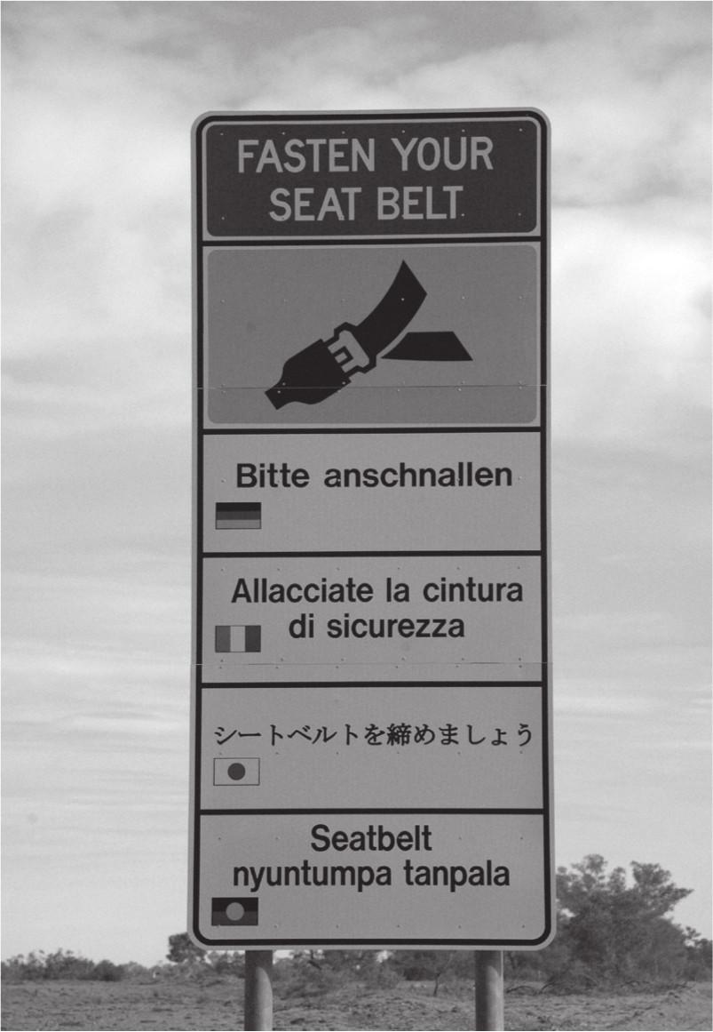 A black and white photograph of a multilingual sign. The top says: fasten your seat belt with a picture of a buckled seat belt beneath it. Four translated lines are included: German, Italian, Japanese and Western Desert.