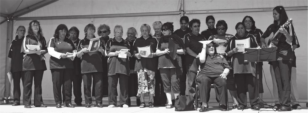 A black and white photograph of a choir made up of middle-aged men and women. They are all standing on a stage, holding a piece of paper. One man is seated and has the paper over his head, to shield his eyes whilst a woman stands to the side, playing a guitar. 
