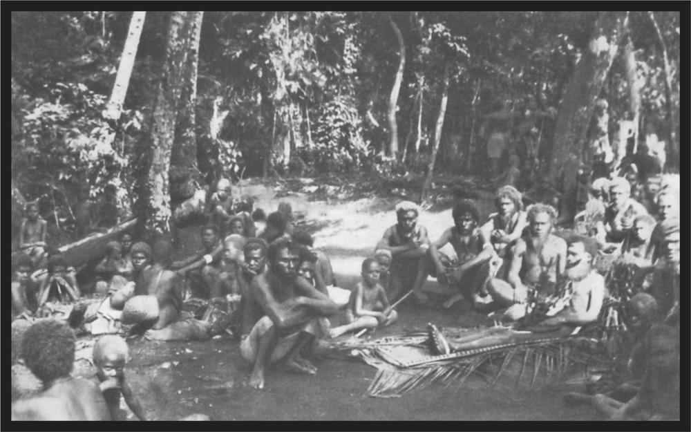 Black and white imageof a large group of men and women sitting on the ground with large trees in the background. There is a corpse sitting on palm fronds.