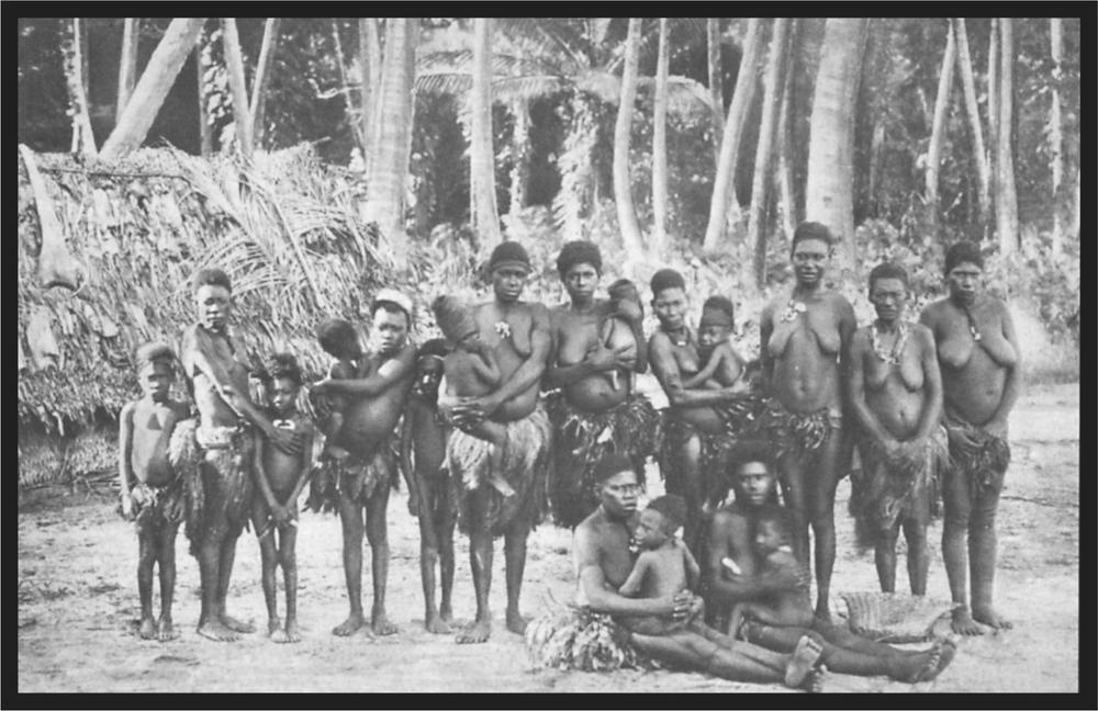 Black and white image of a group of women and chidren standing on the beach in front of large palm trees.