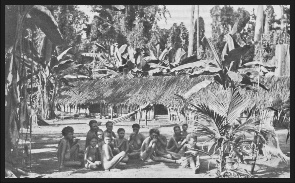 Black and white image of a group of people sitting on the ground of the village in front of a hut.