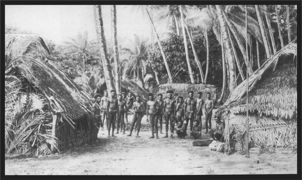 Black and white image of a group of men standing in their village in between numerous huts.