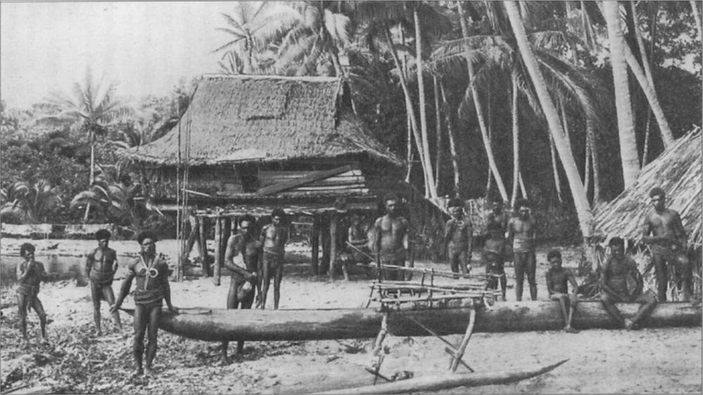 Black and white image of a group of men standing on the beach with long boats and a hut and palm trees in the background.