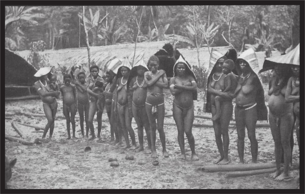 Black and white image of a group of women standing in the sand of the village in front of the huts.