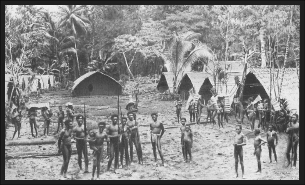 Black and white image of a village, with people standing in the foreground around the village and huts and trees in the background.