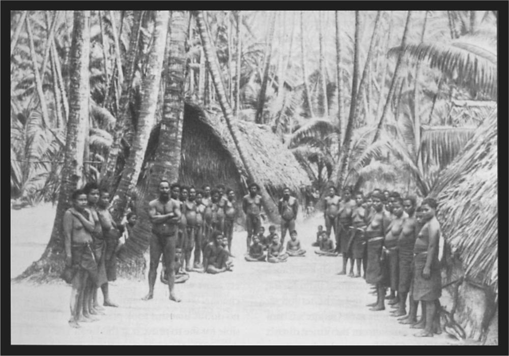 Black and white image of a group standing in the villagewith large palm trees and huts in the background.