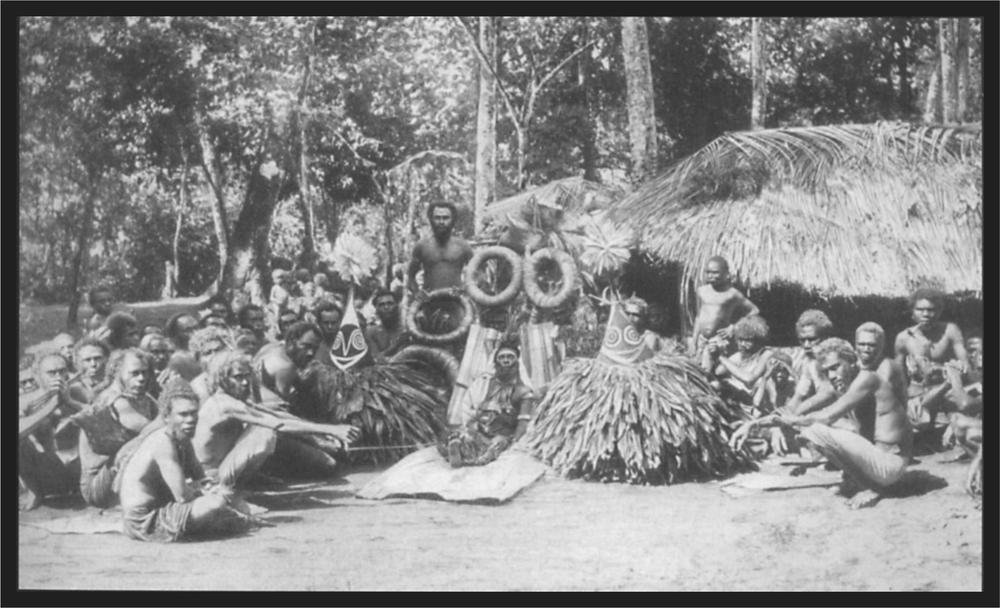 Black and white image of a group of villagers sitting in their village in front of their huts.