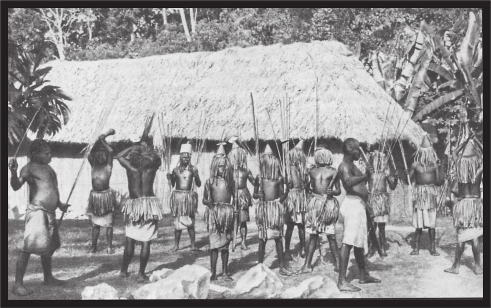 Black and white image of a group of dancers in the village in front of the hut.