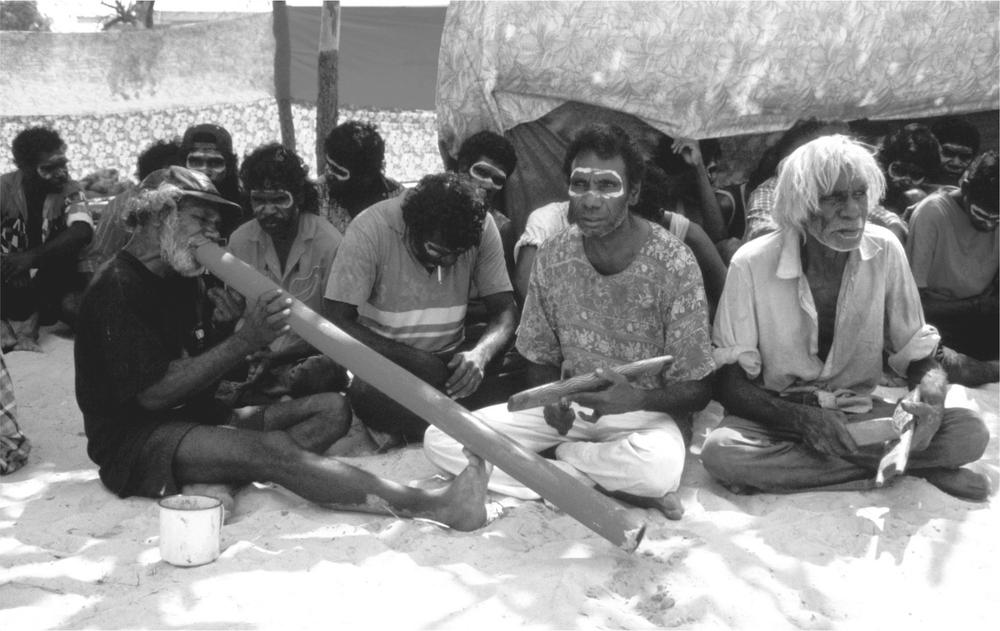 A black and white image of Aboriginal men sitting on the ground and playing the didjeridu at a ceremony.