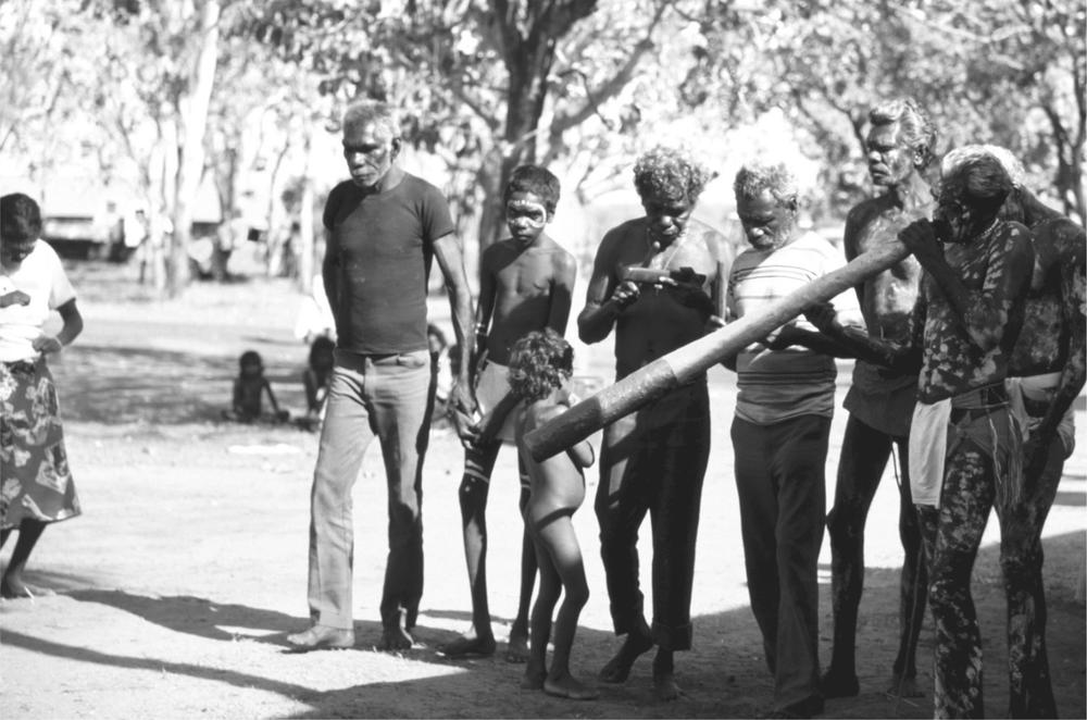 A black and white image of Aboriginal men and a dhild standing. There is one man with his body painted and is playing a didjeridu in the corner of the image.