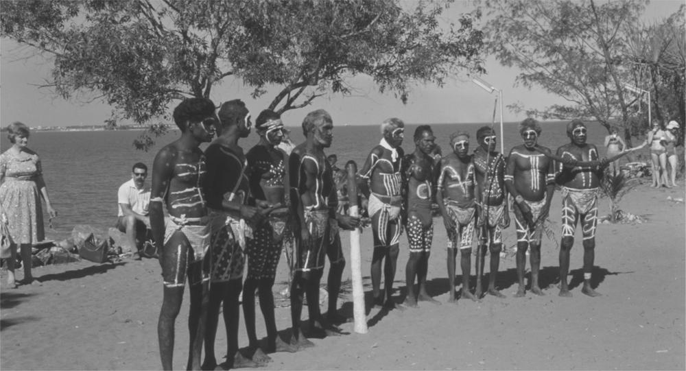 A black and white image of Aboriginal men with body pain standing in a line with a didjeridu.