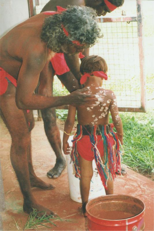 An image of a wangga dancer painting a child dancer. They are wearing a red garmet covering their waists.
