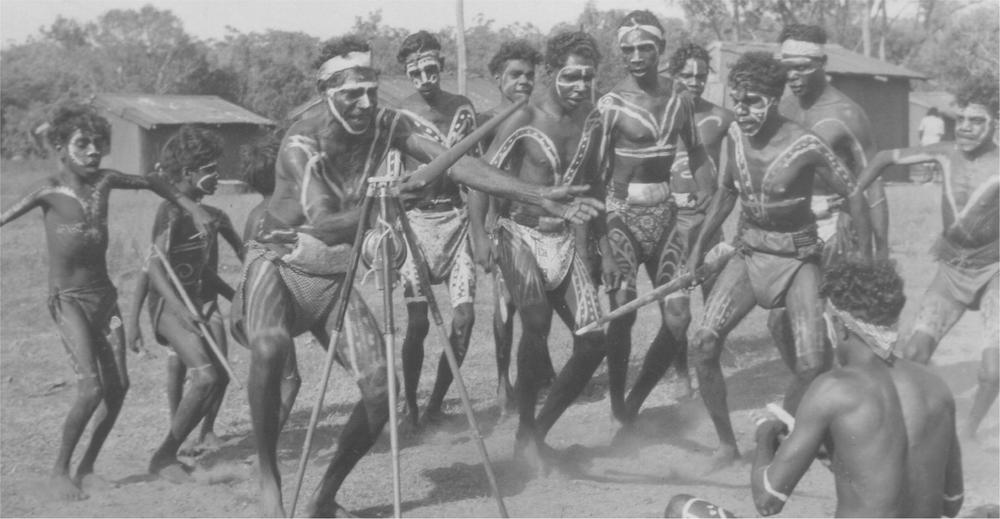 A black and white image of a group of men and children dancing in traditional body paint.