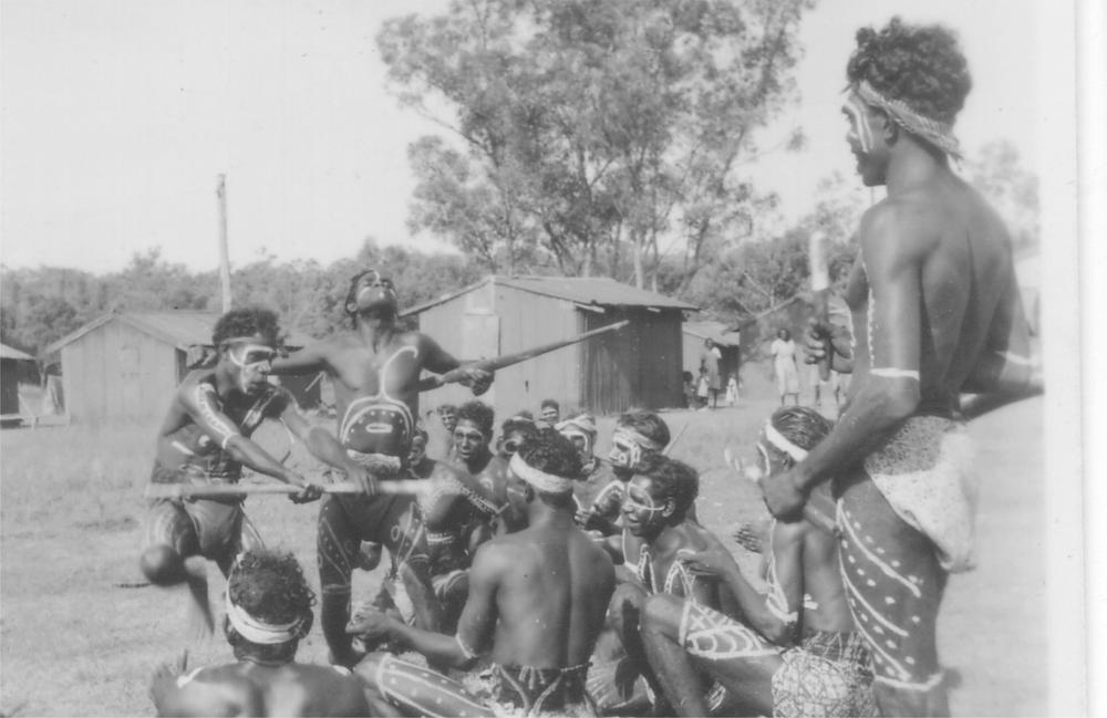 A black and white image of a group of men and boys standing and sitting as they sing and dance.
