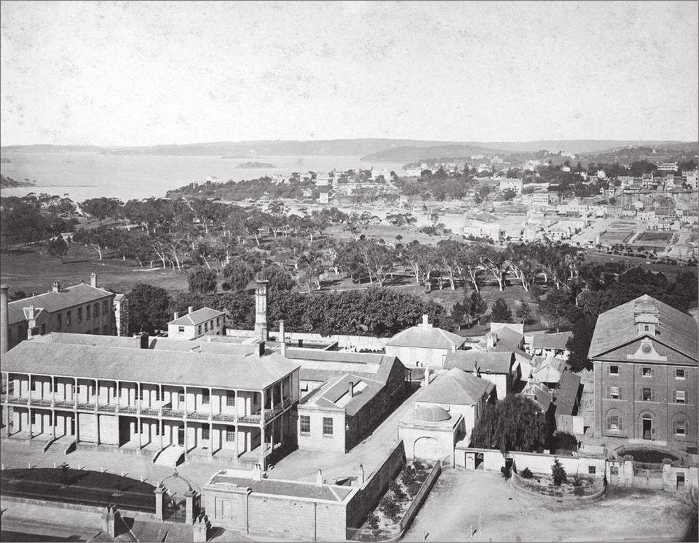 A black and white photograph of the Royal Mint and Hyde Park Barracks. The Domain can be seen behind the barracks along with Woolloomooloo Bay.