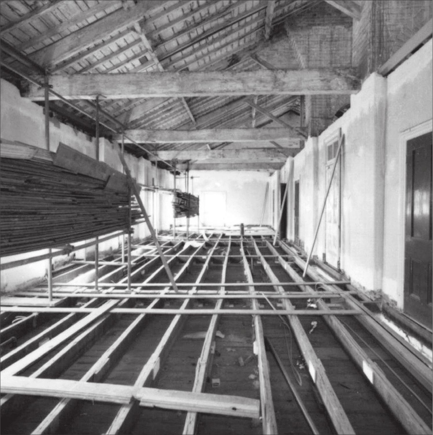 A black and white photograph of a row of criss-crossing planks in an empty room. A stack of floor boards is to the left.