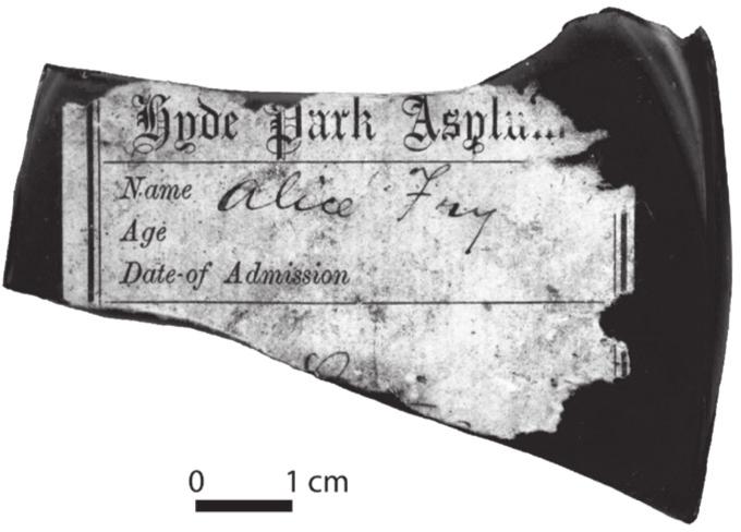 A black and white photograph of a Dispensary Label for Alice Fry on a dark olive-green gin or schnapps bottle. 