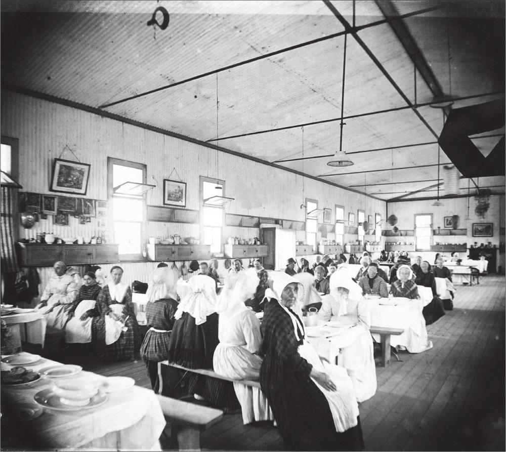 A black and white photograph of a dining room where women are sitting on several rows of tables and benches. They are all wearing a patterned dress with aprons and bonnets. The dining room has a row of open windows and hanging frames.