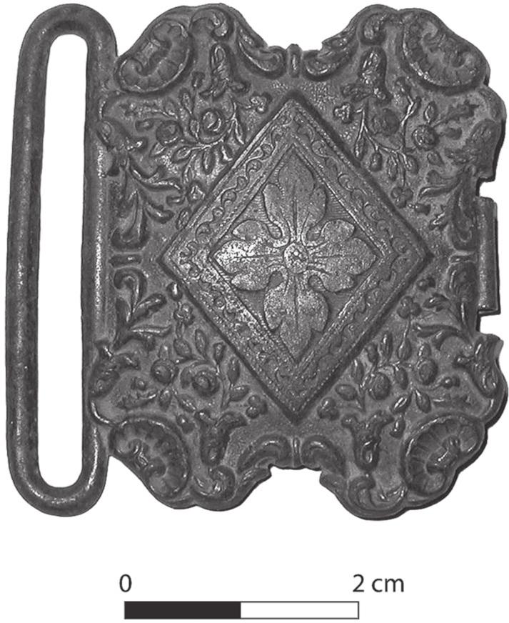 A black and white photograph of a belt buckle with a central lozenge and a four leaf motif. 