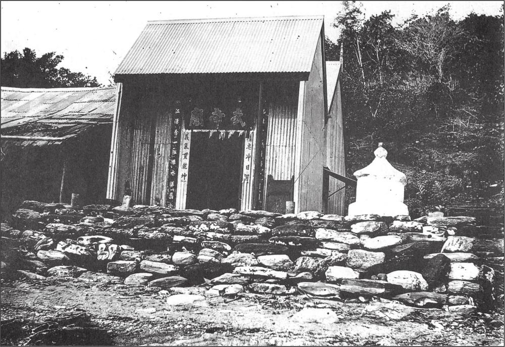 A black and white photograph a fishermen's temple. It has the shape of a small house with a triangular roof but one wall is removed to reveal Chinese banners.