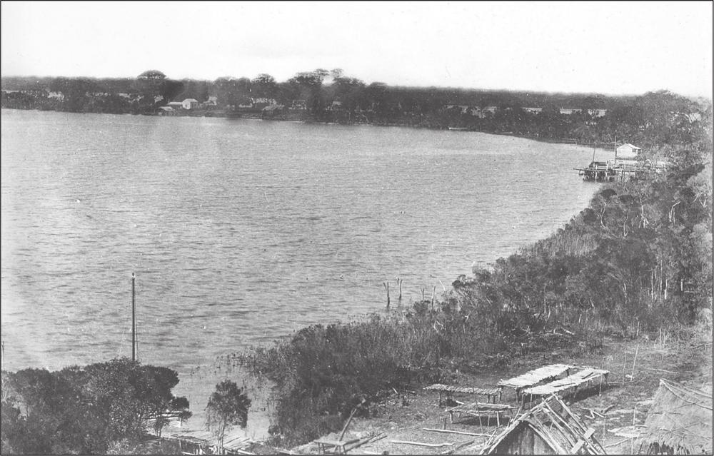 A black and white photograph of a shoreline surrounded by trees. In the bottom right corner of the picture low fish-drying racks are visible.
