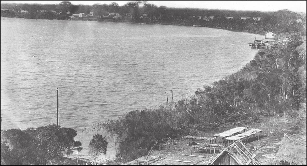 A black and white photograph of a shoreline surrounded by trees. In the bottom right corner of the picture low fish-drying racks are visible.